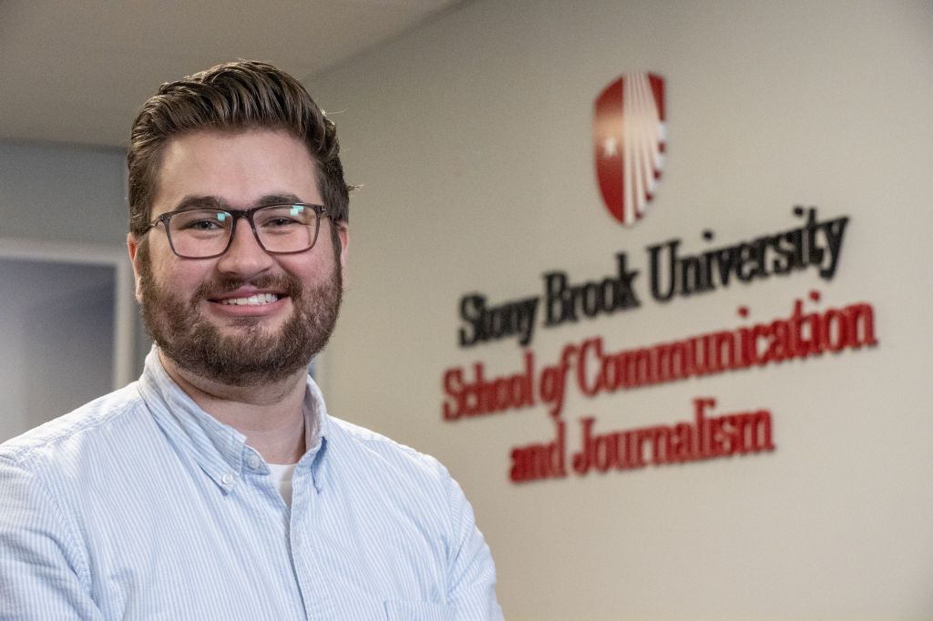 A photo of Matthew Salzano, a white man with brown hair, a beard, and dark glasses, in front of the logo for the Stony Brook University School of Communication and Journalism. 